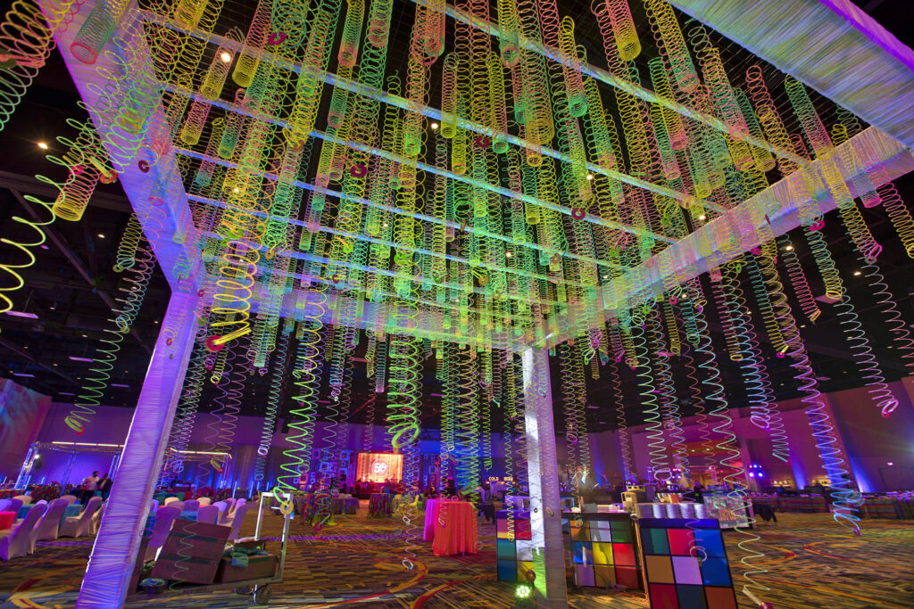 A large event space decorated with hanging colorful Slinkys, bright lights, and retro items like a giant Rubik’s Cube. Round tables with chairs and vibrant decorations create a lively, festive atmosphere.