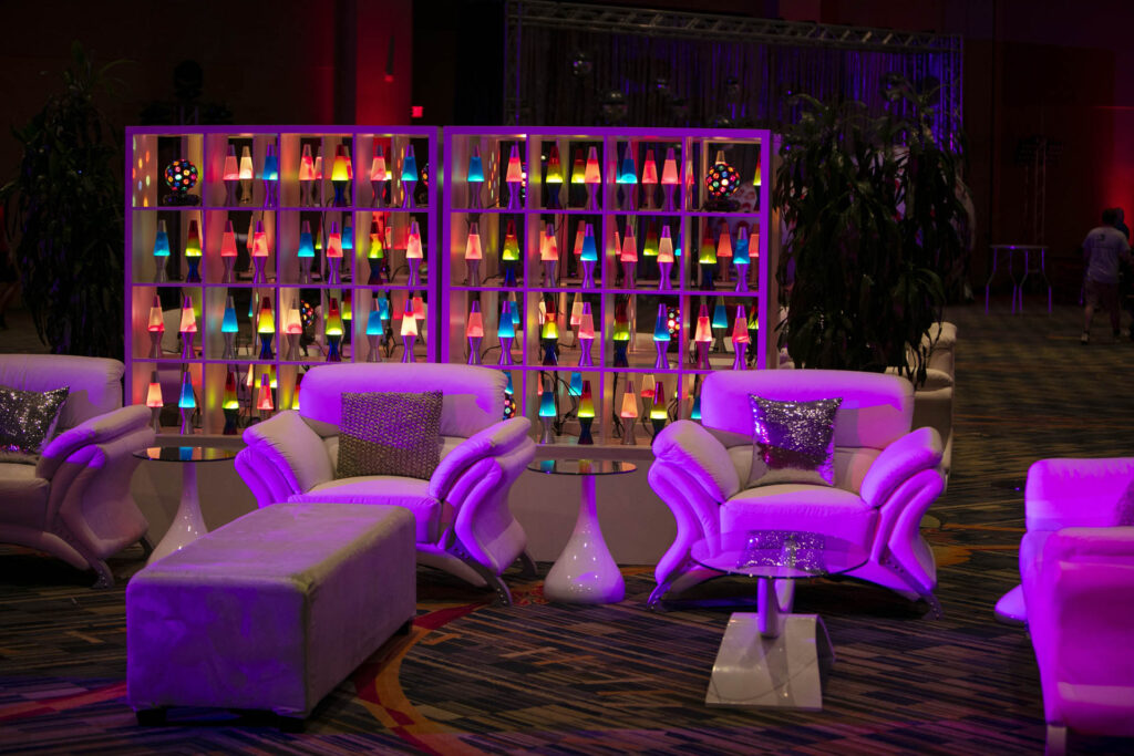 A lounge area with white chairs, a bench, and side tables, all lit by purple lighting. Behind the seating, shelves display many colorful, glowing lava lamps, creating a vibrant and playful atmosphere.