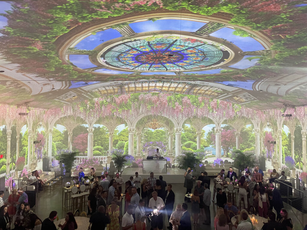 A large crowd gathers indoors under a domed ceiling with a vibrant, garden-themed projection. The ceiling displays a stained glass dome and lush greenery, while guests socialize around tables in an elegant, festive setting.