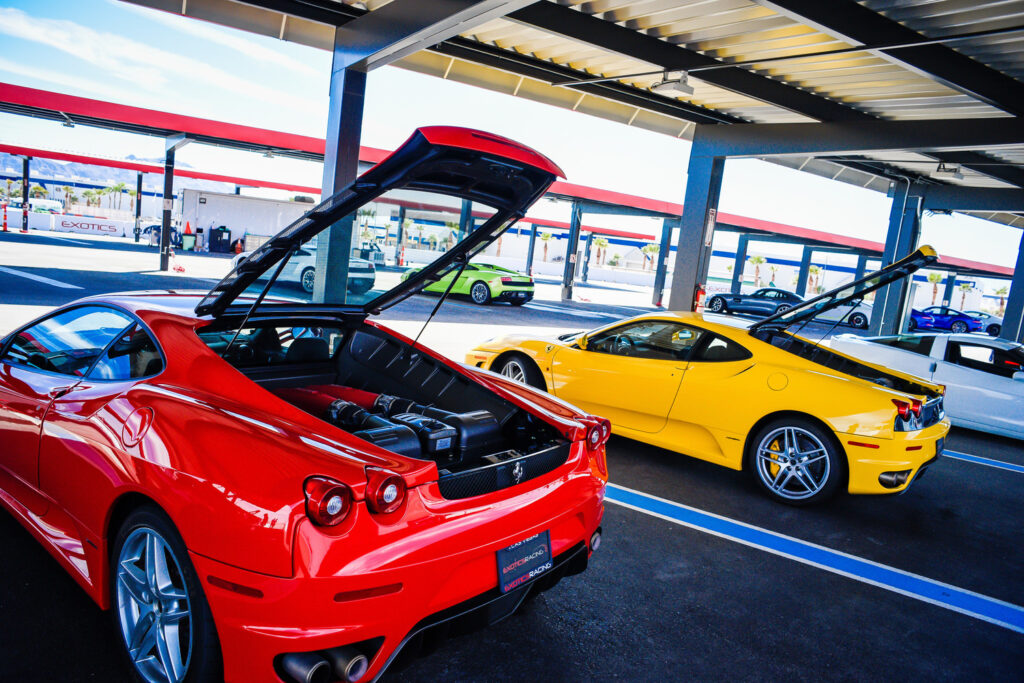 Red and yellow sports cars with their rear engine hoods open are parked side by side under a metal canopy at an outdoor car event, with other cars and people visible in the background.