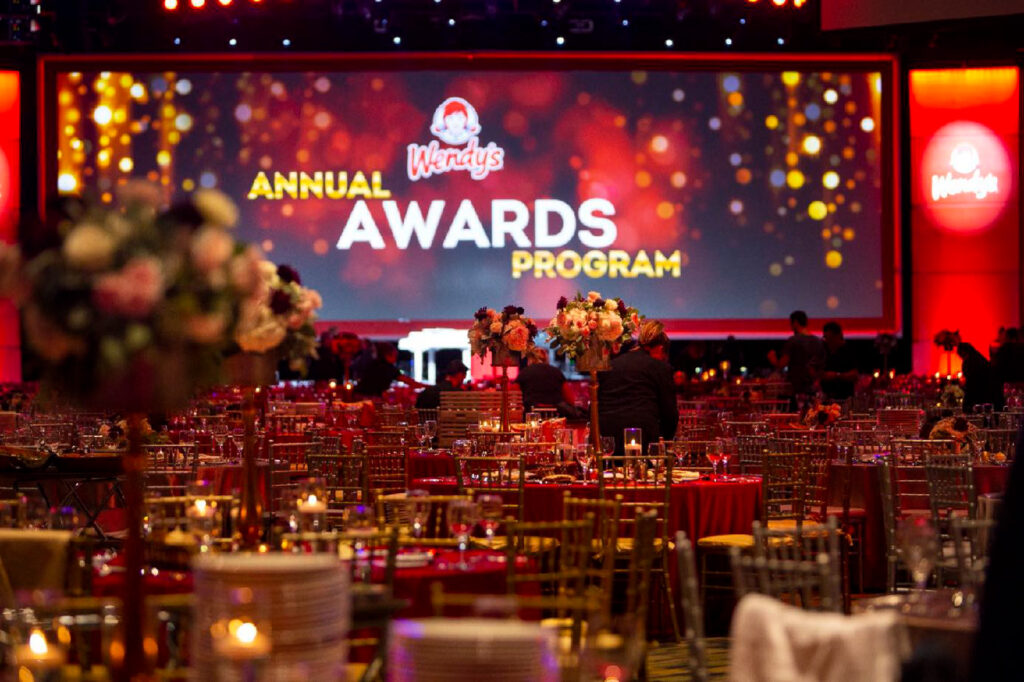 A decorated banquet hall with elegant tables and chairs is set up for Wendy’s Annual Awards Program, with a large illuminated screen displaying the event name in the background.