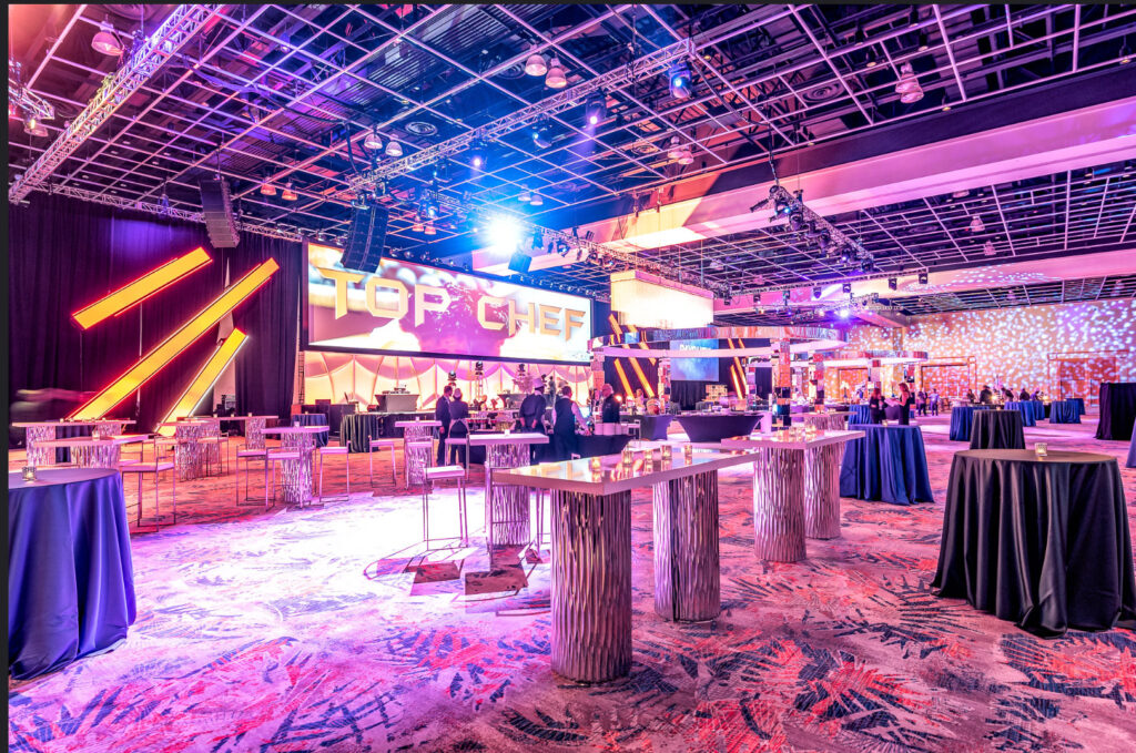 A spacious, modern event venue decorated with high tables and draped linens. Large lit 