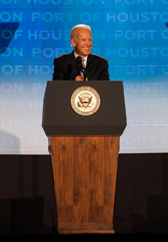 A man in a suit stands and smiles at a podium with the U.S. presidential seal, in front of a blue backdrop that reads 