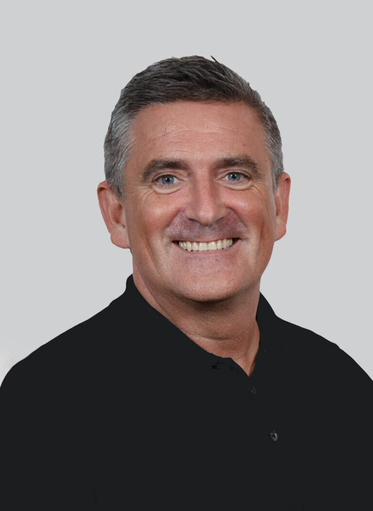 A middle-aged man with short, dark hair smiles at the camera. He is wearing a black collared shirt and is posed against a plain, light gray background.