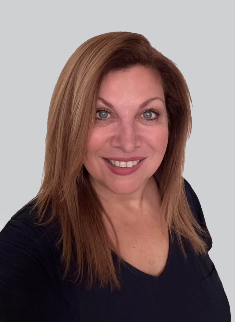 A woman with straight, light brown hair and green eyes smiles at the camera. She is wearing a dark top and is posed against a plain, light gray background.