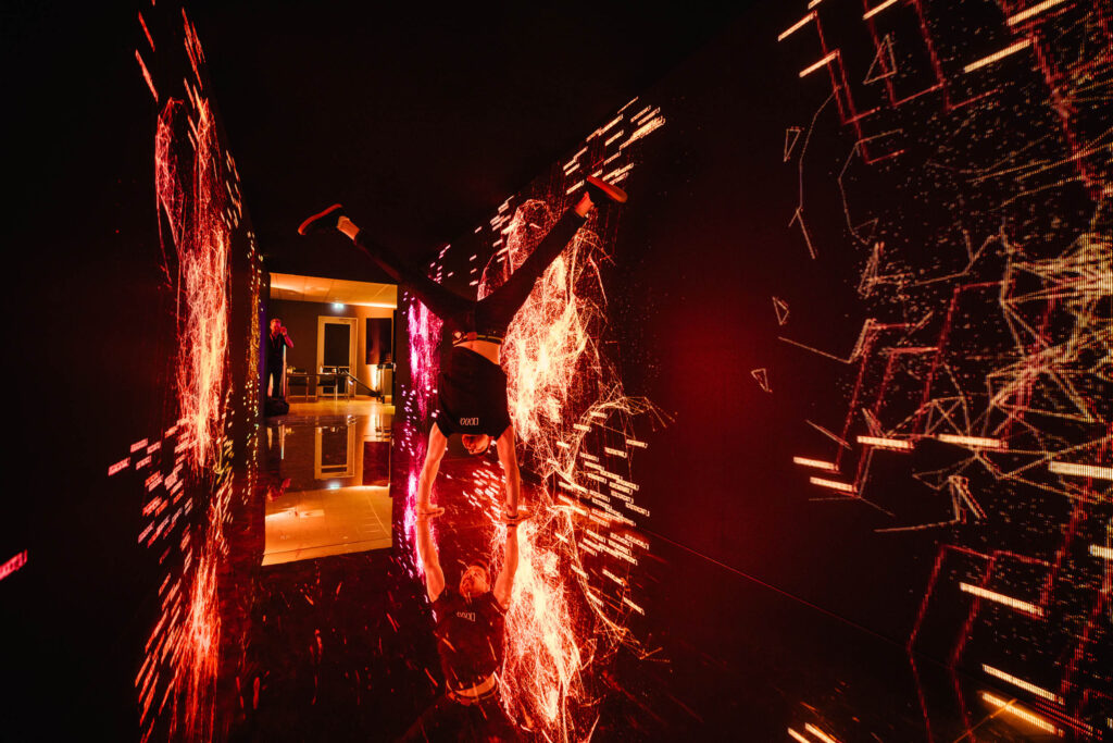 A person performs a handstand in a dark hallway illuminated by vibrant red and orange digital light effects on the walls, creating a futuristic, energetic atmosphere. Their reflection is visible on the glossy floor.