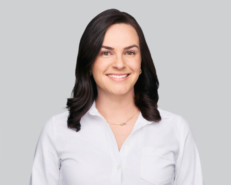 A woman with dark, shoulder-length hair wearing a white button-up shirt and a delicate necklace smiles at the camera against a plain light grey background.