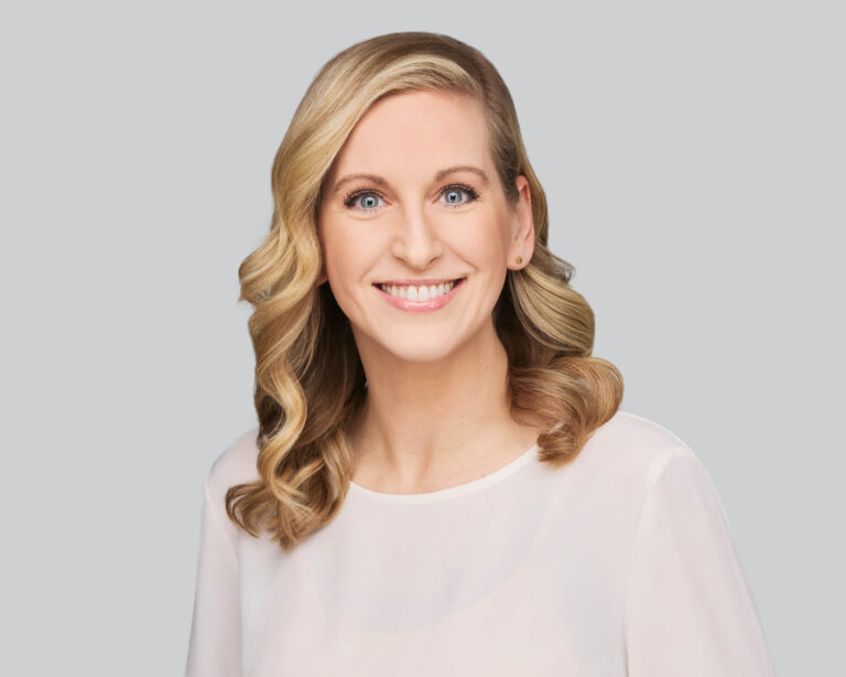 Smiling woman with long, wavy blonde hair wearing a sheer white blouse, posed against a plain light gray background.