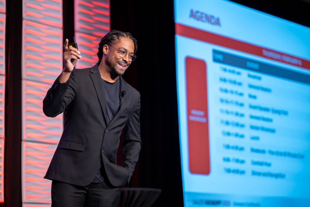 A man in a black suit and glasses smiles while speaking on stage, holding a marker. A large screen beside him displays an agenda with times and event titles in a conference setting.