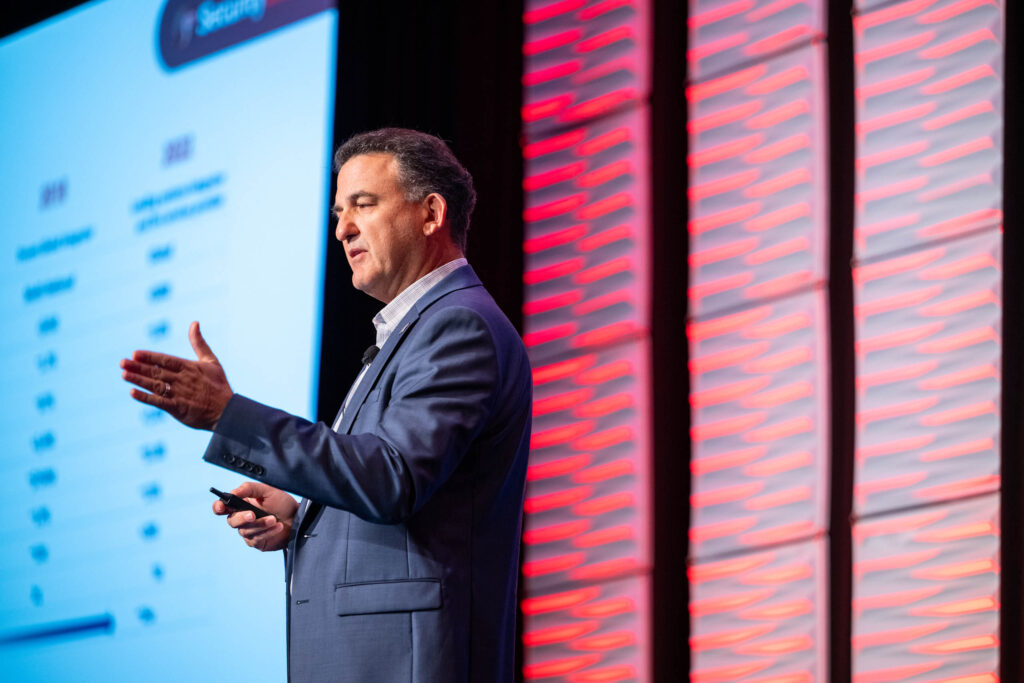 A man in a suit speaks on stage, gesturing with one hand, in front of a screen displaying charts and text, with a red patterned wall in the background.
