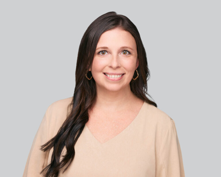 A woman with long dark hair and light skin smiles at the camera. She wears a light beige top and gold hoop earrings, standing against a plain light gray background.