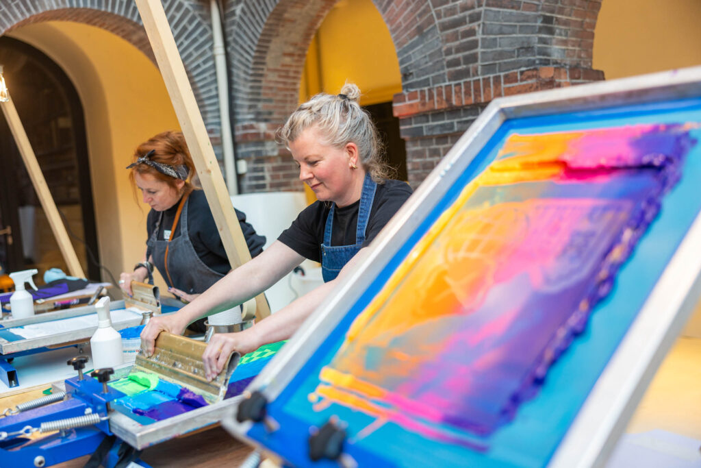 Two women wearing aprons work on colorful screen printing projects at a table, with vibrant inks on a screen in the foreground, in a room with arched brick walls.