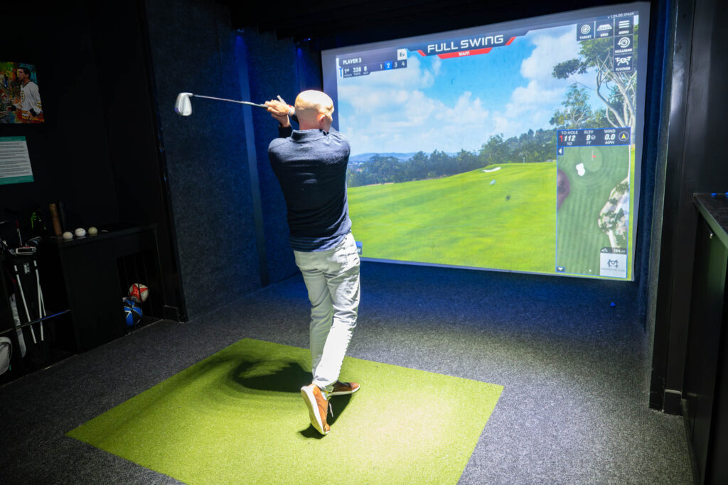 A man swings a golf club indoors on an artificial turf mat, facing a large screen displaying a virtual golf course simulation labeled “FULL SWING.” Golf equipment is visible to the side.