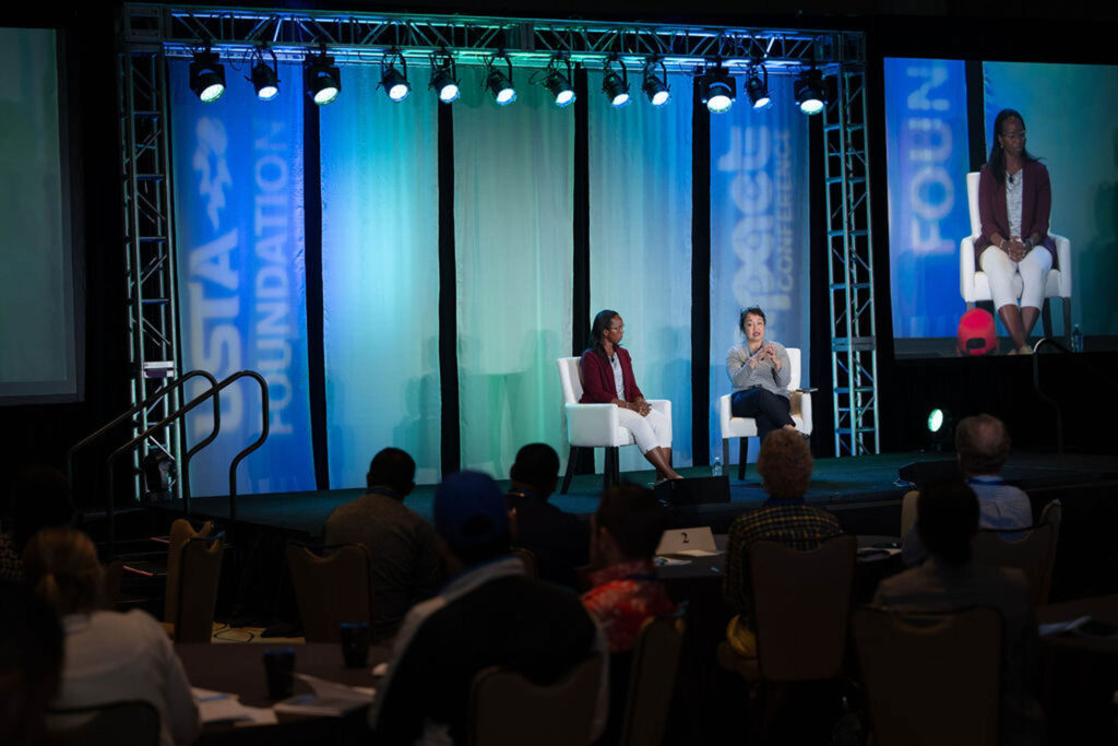 Two women sit on stage having a discussion in front of an audience. The stage is lit with blue and green lights, and banners reading 