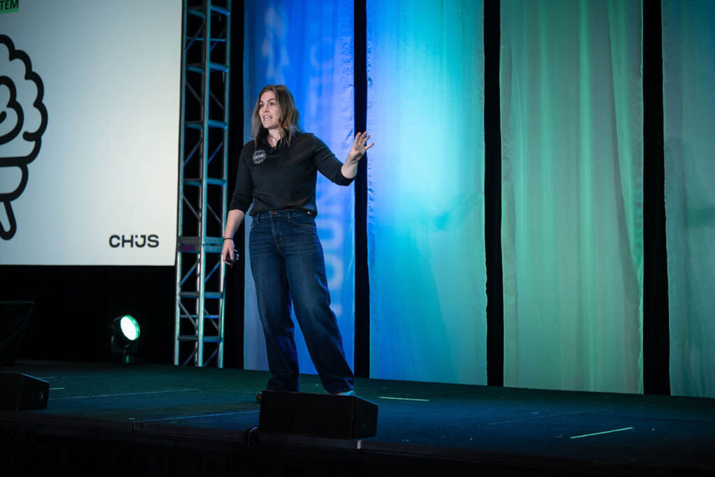 A person stands and gestures on a stage with blue and green curtains behind them. A spotlight shines from the left, and a large screen displays part of a graphic and the text “CHJS.”.