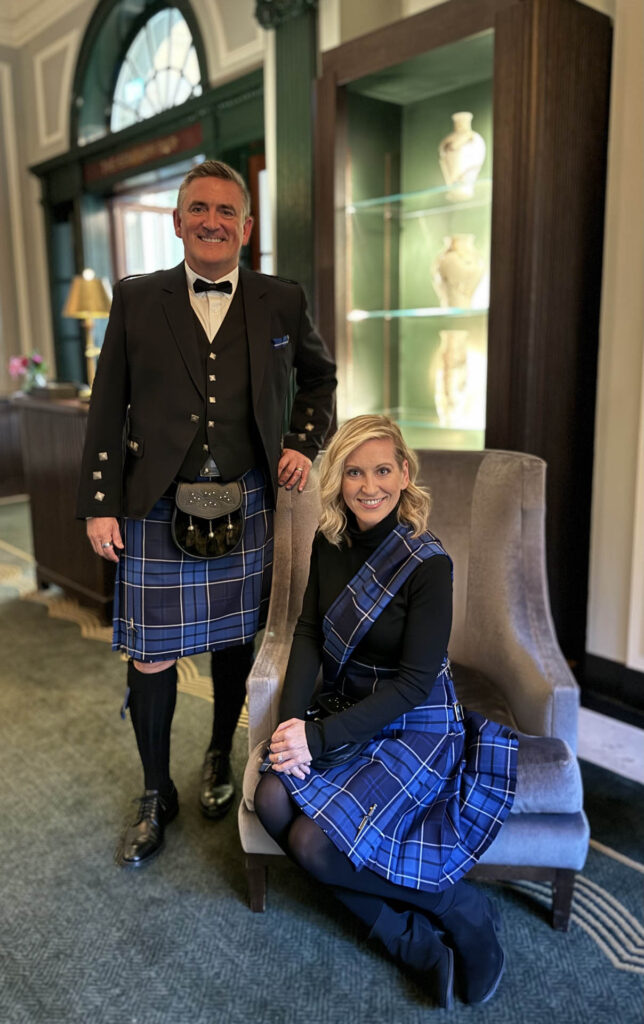 A man and woman dressed in matching blue tartan kilts pose indoors; the man stands beside a chair while the woman sits on it, both smiling in a refined, elegant setting with decorative vases behind them.