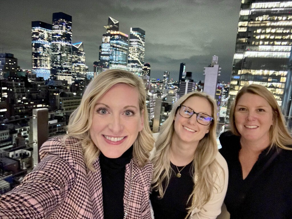 Three women smiling and posing for a selfie on a rooftop at night, with a city skyline of tall, illuminated buildings in the background.