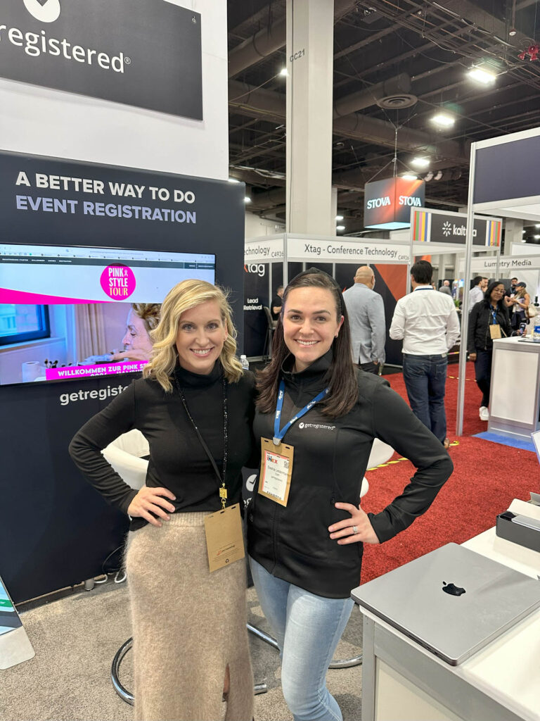 Two women smiling and standing together at a trade show booth for an event registration company. One woman wears a black top and beige skirt, the other wears a black jacket and blue lanyard. A laptop and expo displays are visible.