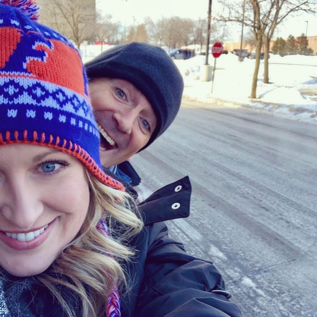 A smiling woman in a colorful winter hat takes a selfie outdoors with a man playfully photobombing behind her. Both are bundled up in warm coats on a snowy street with bare trees in the background.