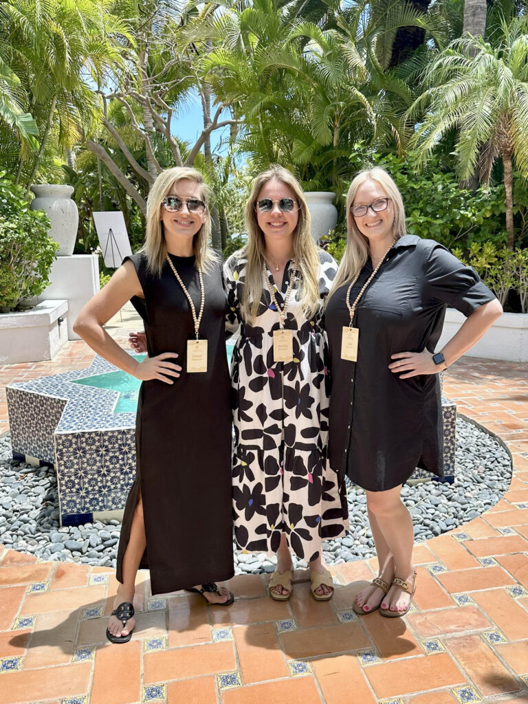 Three women stand smiling outdoors in front of a decorative tiled fountain, wearing sunglasses, lanyards, and summer dresses. Lush green plants and palm trees surround them under bright sunlight.