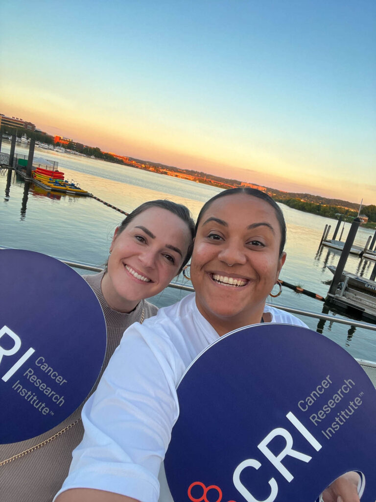 Two smiling women hold large round Cancer Research Institute signs while standing on a dock by calm water at sunset, with boats and a distant shoreline in the background.