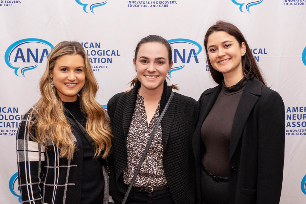 Three women smiling and standing together in front of an American Neurological Association backdrop at an event.