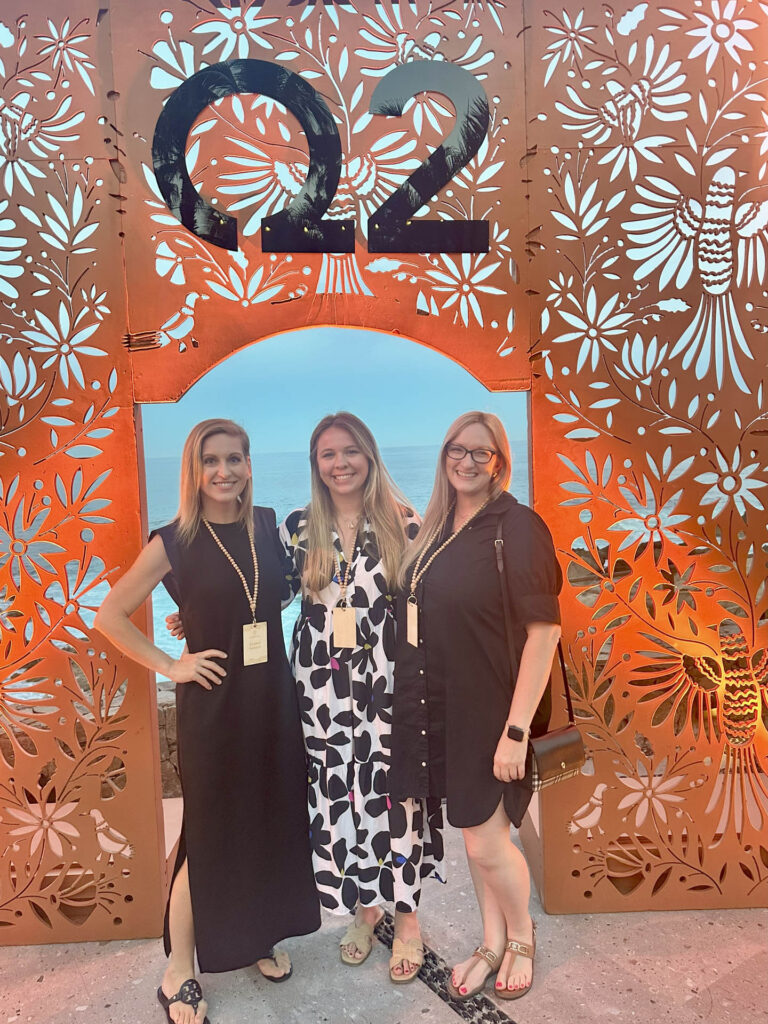 Three women stand smiling in front of an ornate, patterned orange backdrop with “Q2” displayed at the top. The ocean is visible through a central archway. All three wear dresses, event lanyards, and sandals.