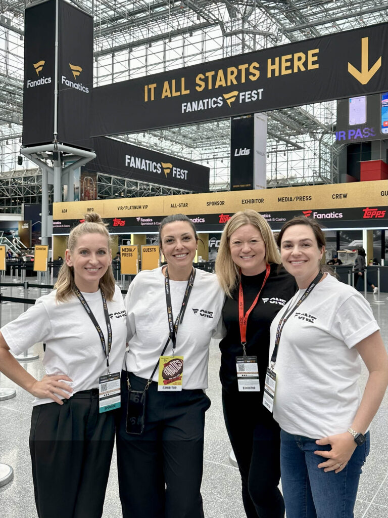 Four smiling women pose together in matching white shirts at Fanatics Fest. Large signs and banners with “It All Starts Here” and “Fanatics Fest” hang in a spacious convention center behind them.