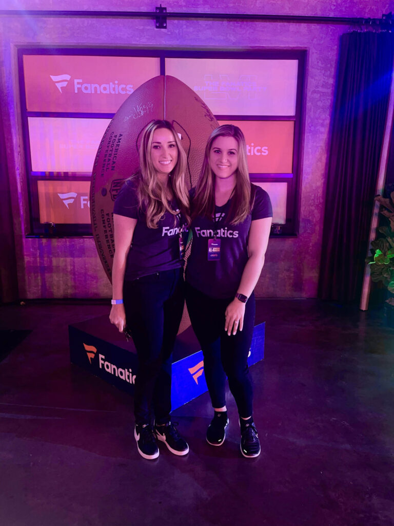 Two women wearing matching Fanatics shirts and dark pants stand smiling in front of a large football prop and Fanatics signs in a purple-lit indoor setting.
