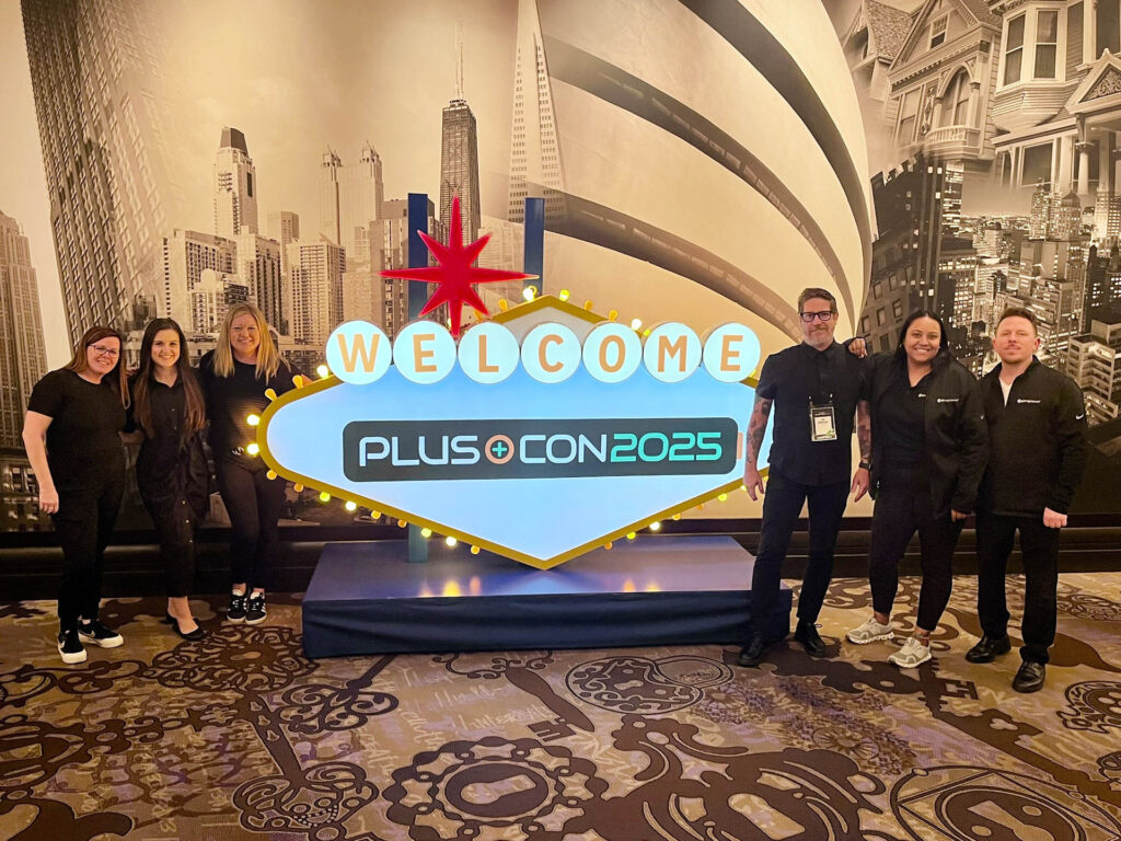 Six people stand smiling beside a large, colorful sign that reads “Welcome PLUS+CON 2025” in a room decorated with black-and-white cityscape murals and patterned carpet.