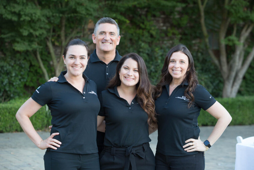 Four people, three women and one man, stand closely together outdoors, smiling at the camera. They all wear matching black polo shirts. Greenery and a paved area are visible in the background.