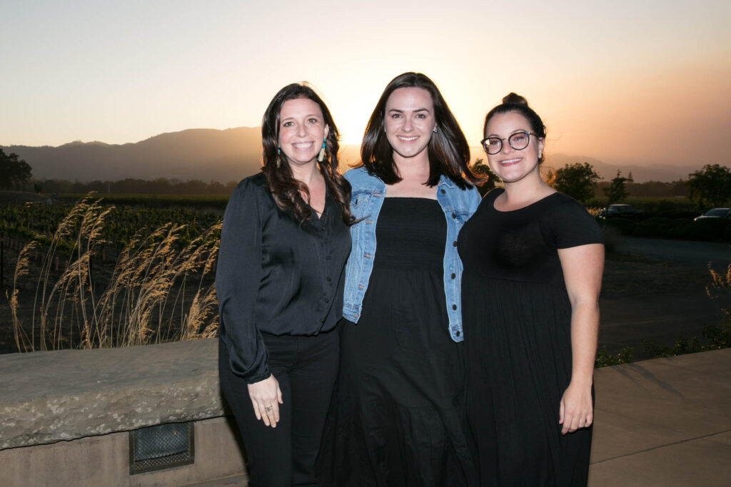 Three women stand close together outdoors at sunset, smiling at the camera. Behind them are grassy fields, a stone wall, and distant mountains under a bright sky. They all wear black outfits, with one in a denim jacket.
