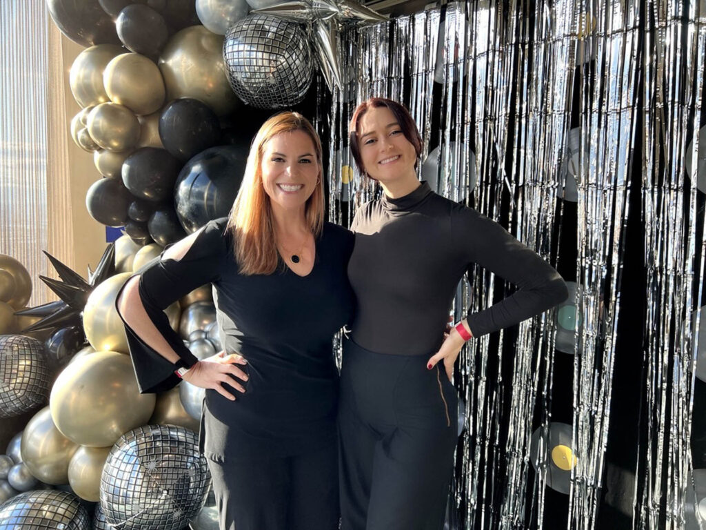 Two women in black outfits stand smiling in front of metallic balloon decorations and silver tinsel streamers, with sunlight shining in from the side.