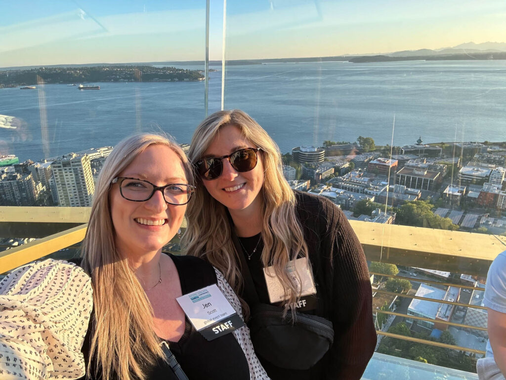 Two women with long blonde hair wearing glasses and staff badges smile for a selfie on an outdoor observation deck overlooking a city, water, and distant mountains at sunset.