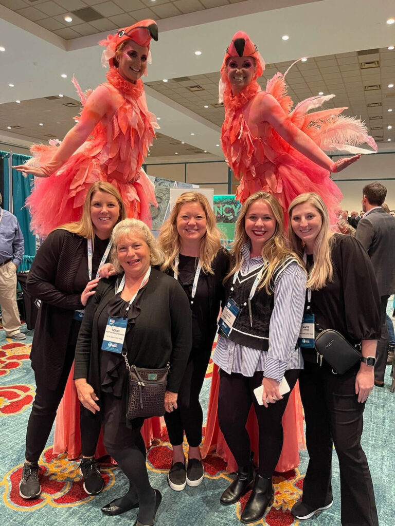 Five smiling women pose together at an indoor event, while two performers on stilts dressed in pink flamingo costumes stand behind them, also smiling. The background shows booths and other attendees.