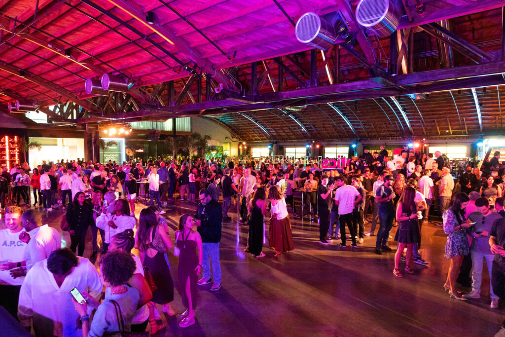 A large crowd of people socializes and mingles at an indoor event under a wooden, vaulted ceiling with colorful lighting. Some people stand in groups, while others walk around the spacious venue.
