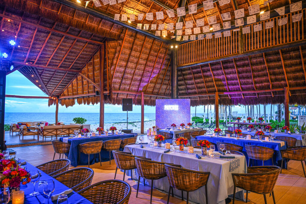 A vibrant beachside venue with thatched roof, decorated tables with flowers, blue tablecloths, and hanging papel picado banners, overlooking the ocean at sunset.