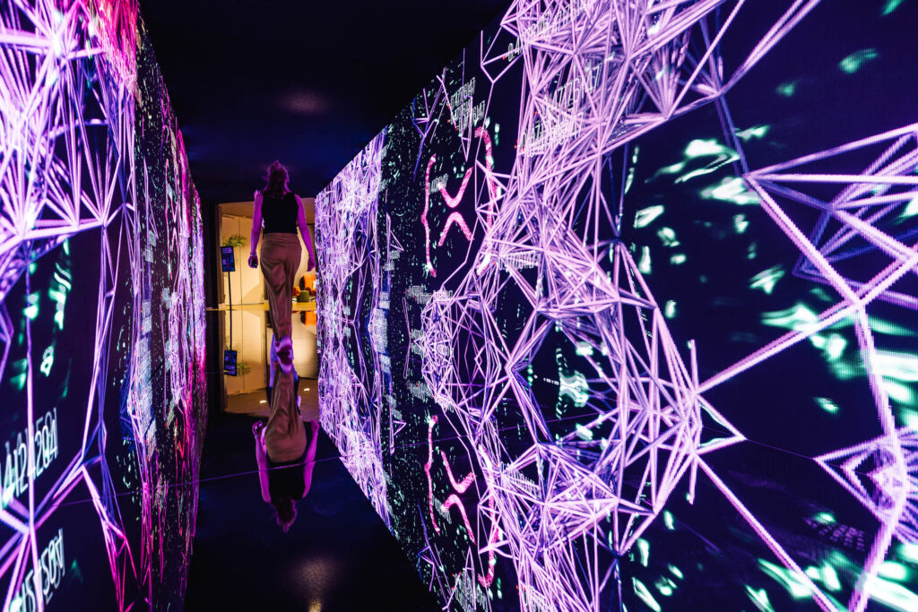 A person walks through a tunnel with vibrant digital art on the walls, featuring glowing, geometric purple and pink patterns and reflections, creating an immersive, futuristic atmosphere.