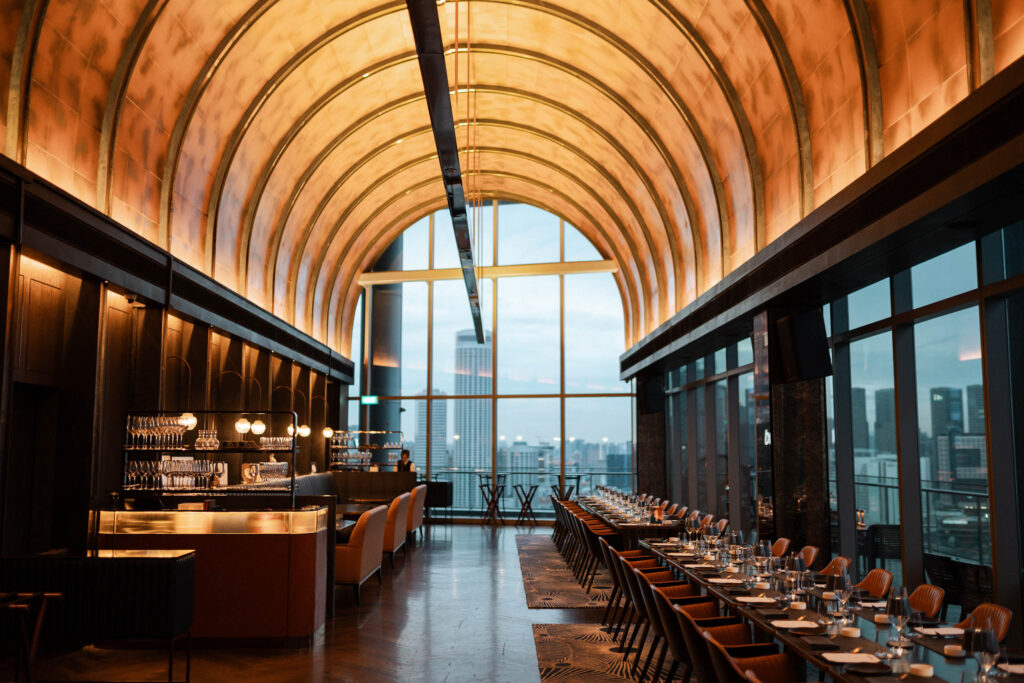 Elegant restaurant interior with a high, golden arched ceiling, floor-to-ceiling windows showing a city skyline, a bar area on the left, and long tables set for dining on the right.