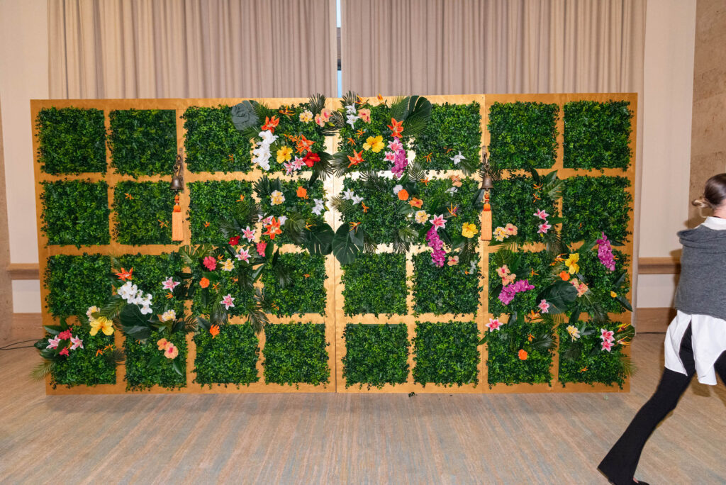 A green wall panel decorated with colorful flowers is set indoors against beige curtains; a person in a gray sweater and white shirt walks past on the right side.