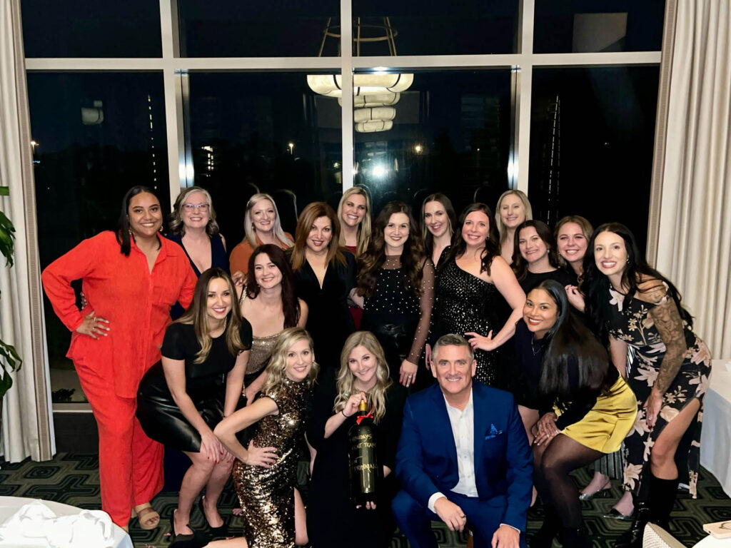 A group of 18 adults, mostly women dressed in evening attire, pose and smile together indoors in front of large windows at night. One woman in front holds a large trophy.