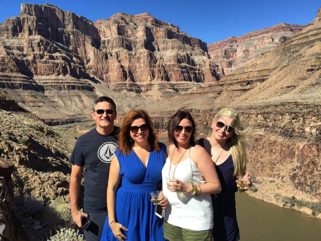 Four people stand smiling in front of a scenic canyon landscape with rocky cliffs and a river below, holding drinks and enjoying a sunny day.