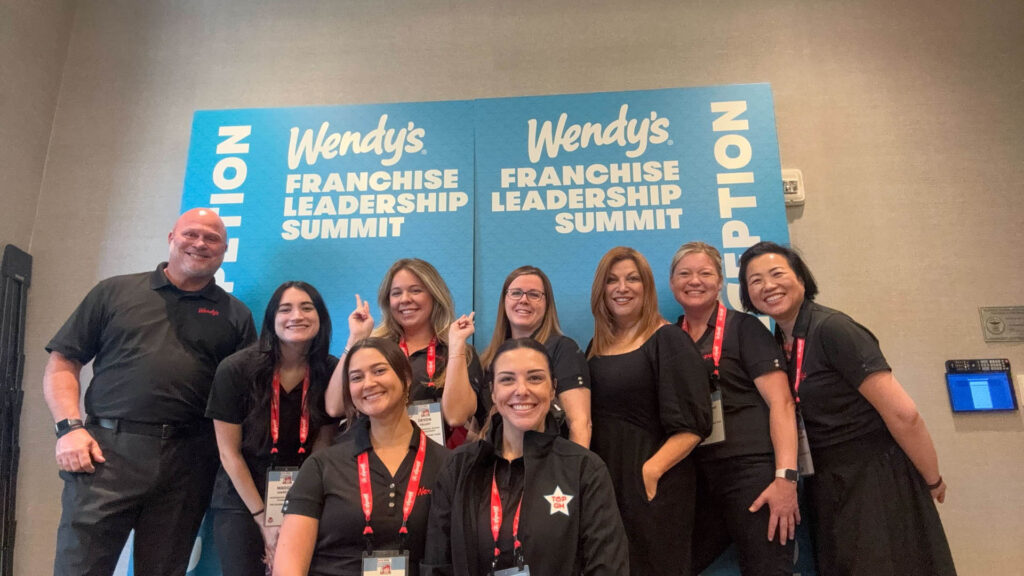 A group of nine people in black shirts pose and smile in front of blue Wendy's Franchise Leadership Summit signs at a conference. Several are wearing red lanyards and name badges.