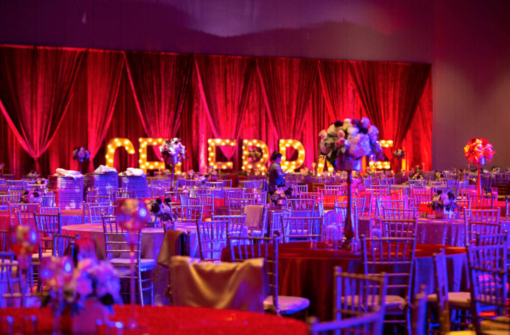 A banquet hall decorated with red and purple lighting, round tables with floral centerpieces, silver chairs, and a large illuminated “CELEBRATE” sign against a red curtain backdrop.
