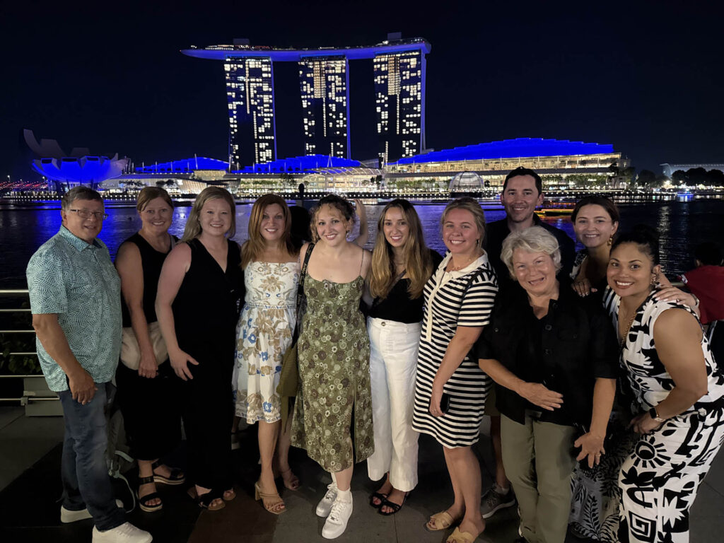 A group of eleven people smile for a photo at night in front of the illuminated Marina Bay Sands hotel in Singapore, with city lights reflecting on the water behind them.