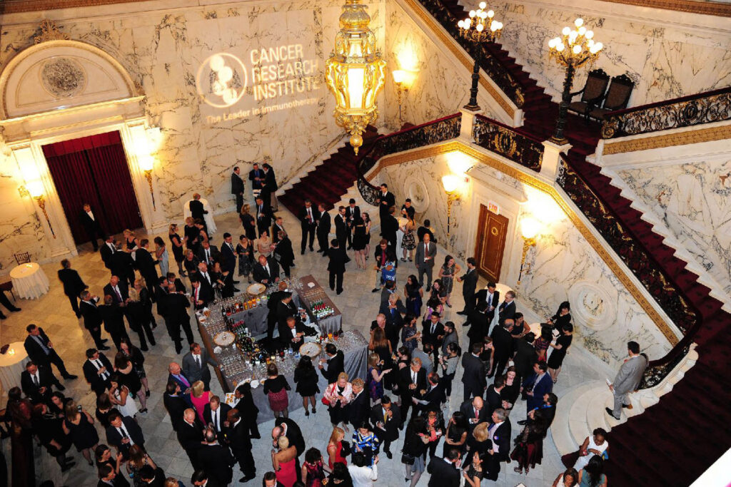 A large group of people in formal attire gather in a grand marble hall with a staircase, ornate chandeliers, and a “Cancer Research Institute” logo projected on the wall. Guests mingle around tables with drinks.