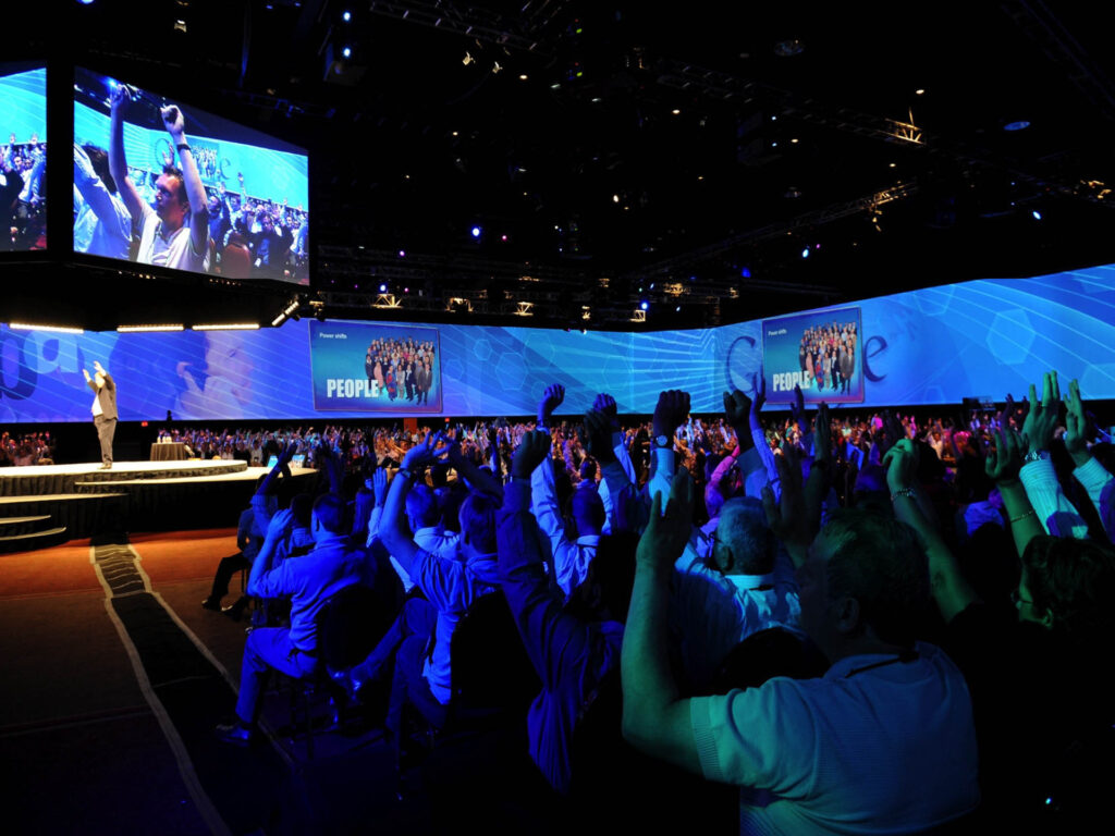 A large audience sits in a darkened conference hall with blue lighting, raising their hands toward a stage where a speaker stands. Multiple large screens display images of the crowd and presentation slides.